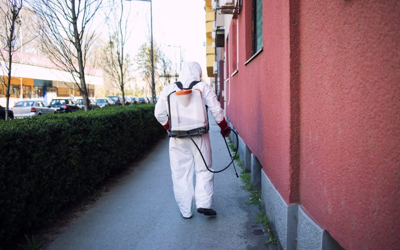 Unrecognizable worker in chemical protection suit spraying disinfectant on public surfaces.